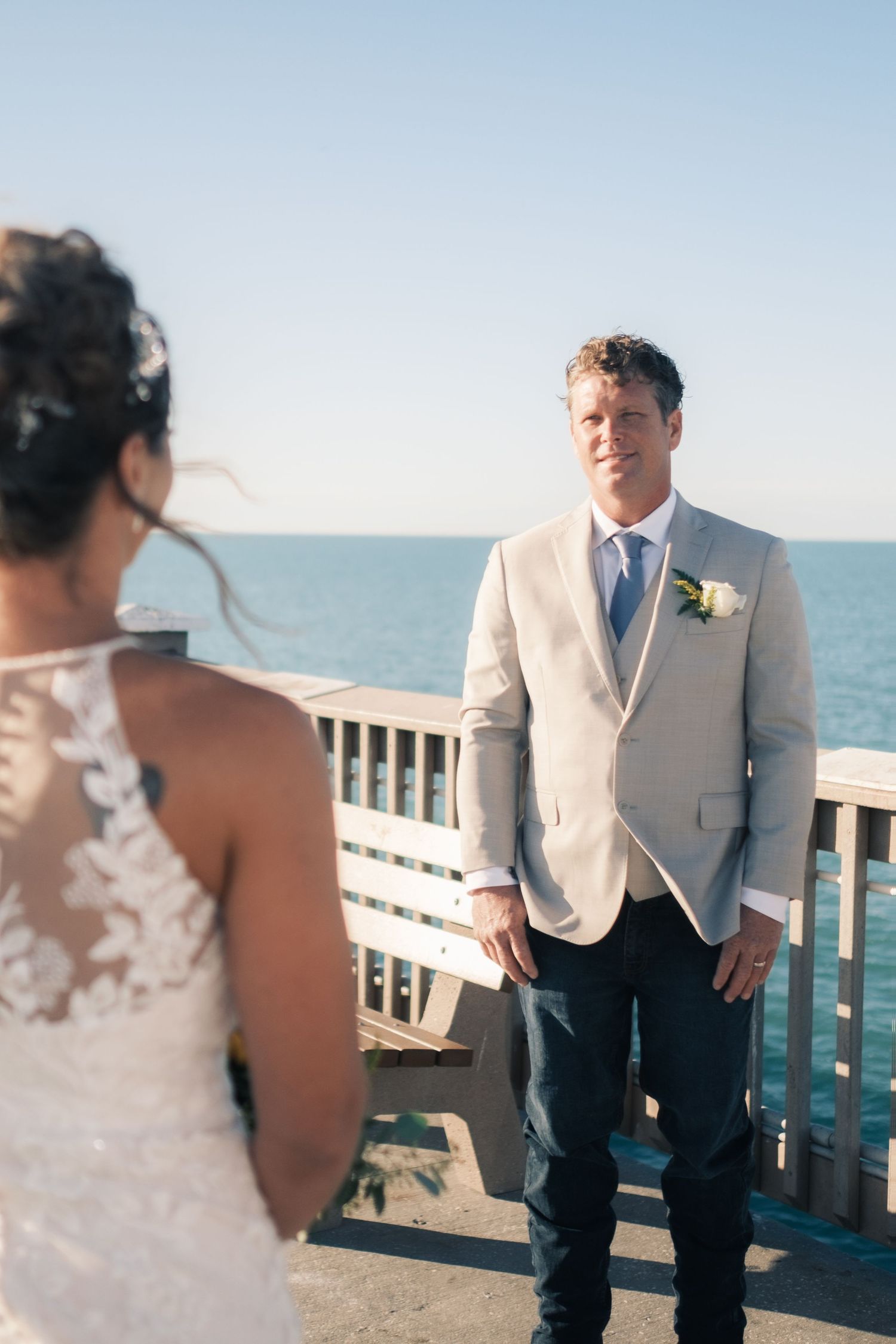 Groom in tan suit watches bride approach during first look on a seaside deck.