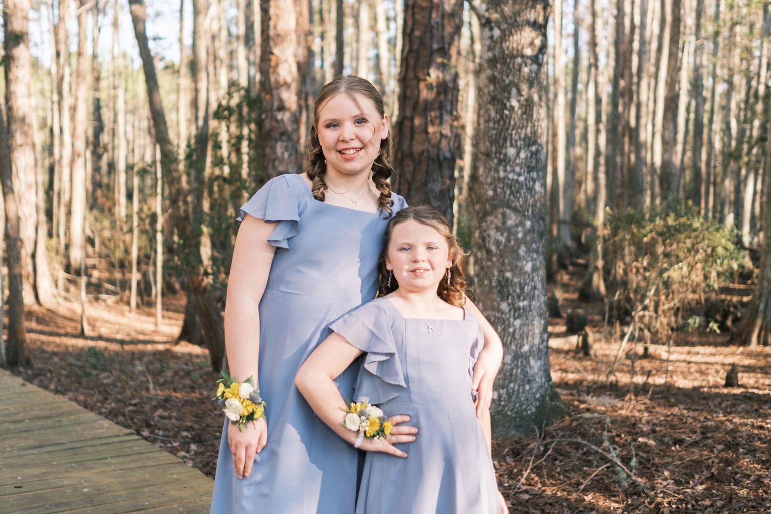 Two bridesmaids in matching blue dresses with wrist corsages pose together in a sunlit pine forest.