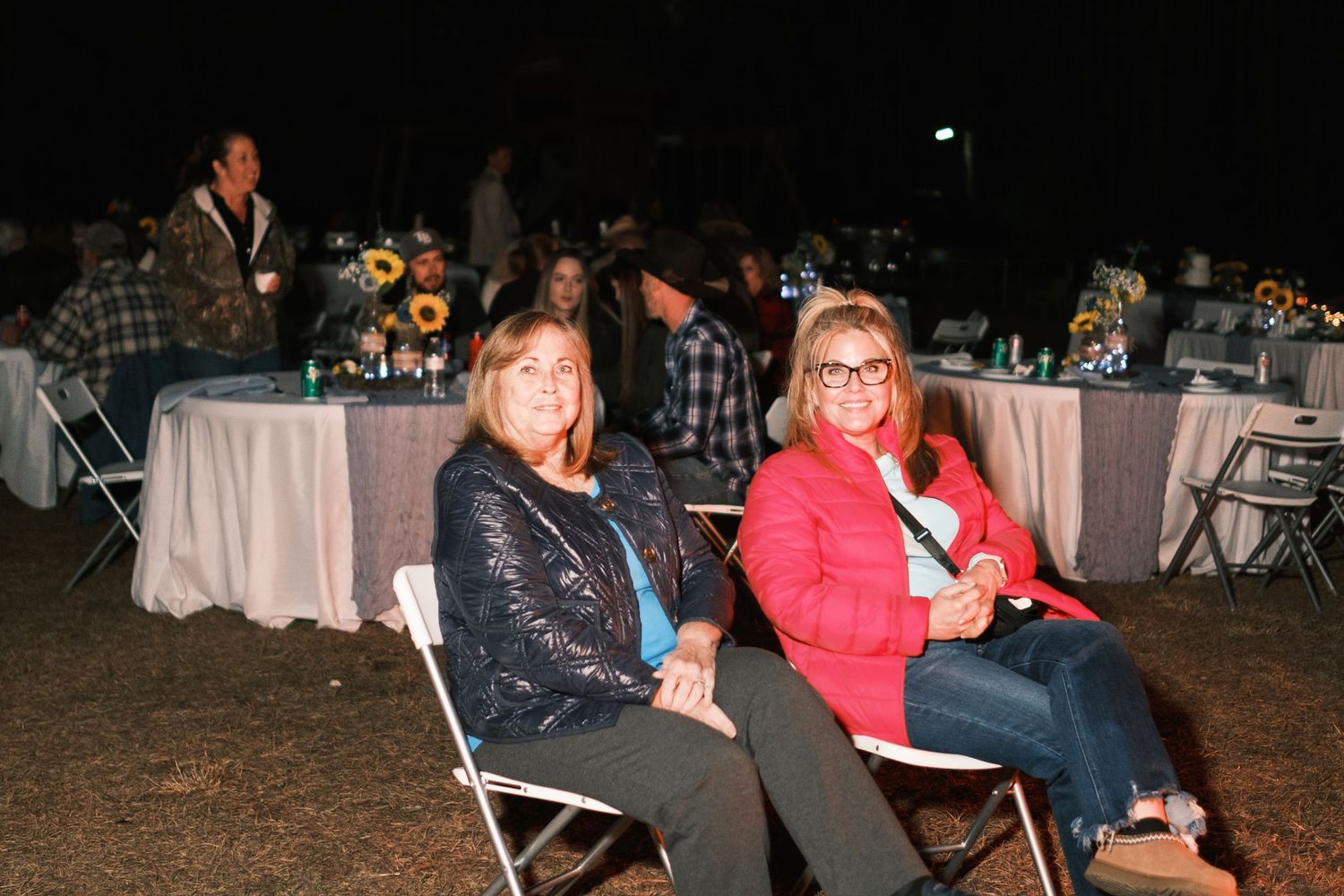 Two women smile while seated at an outdoor evening wedding reception with sunflower table arrangements.