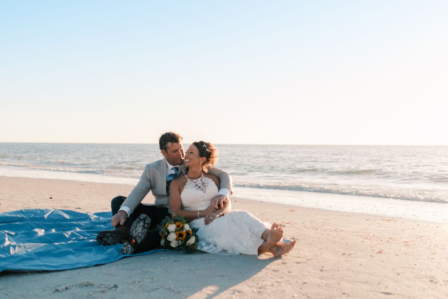 Bride and groom sit together on blanket at beach during golden hour sunset.