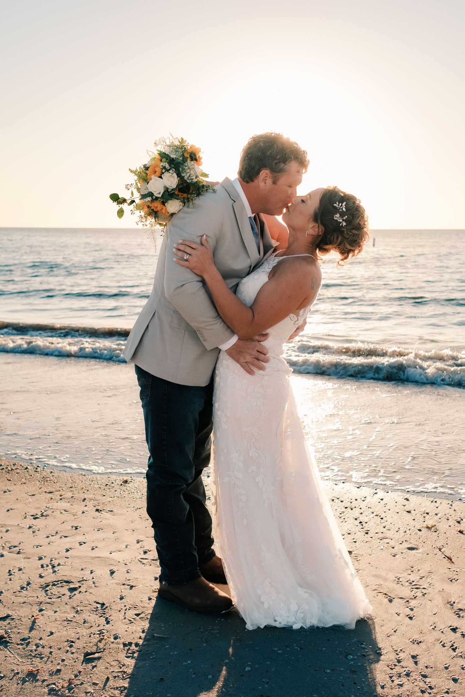 Bride and groom share a kiss on the beach at sunset holding floral bouquet.