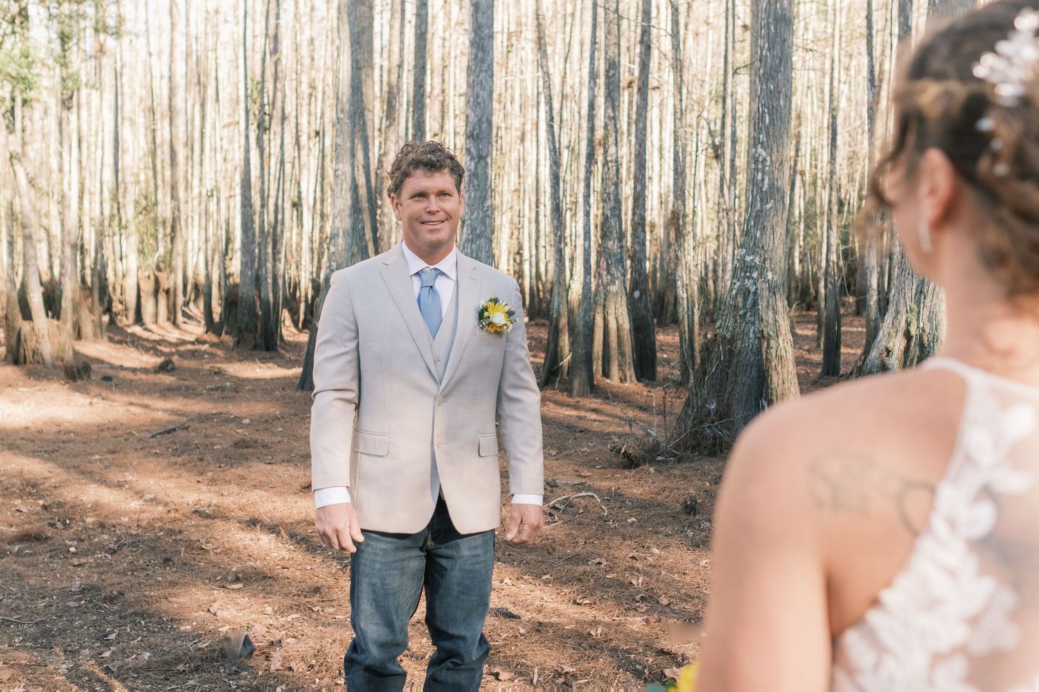 Groom in gray suit smiles at bride during outdoor woodland first look wedding moment.
