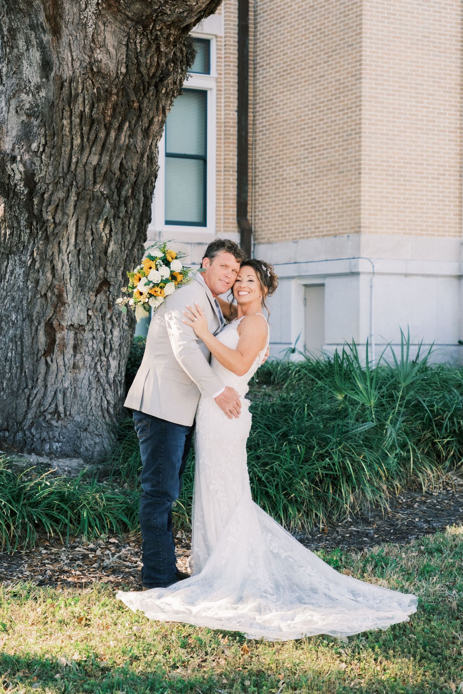 Groom embraces bride from behind near a large tree, bride holds yellow and white bouquet, both smiling.