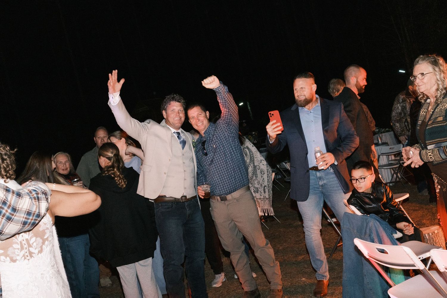 Wedding guests celebrating and dancing outdoors at night around a bonfire at a reception.