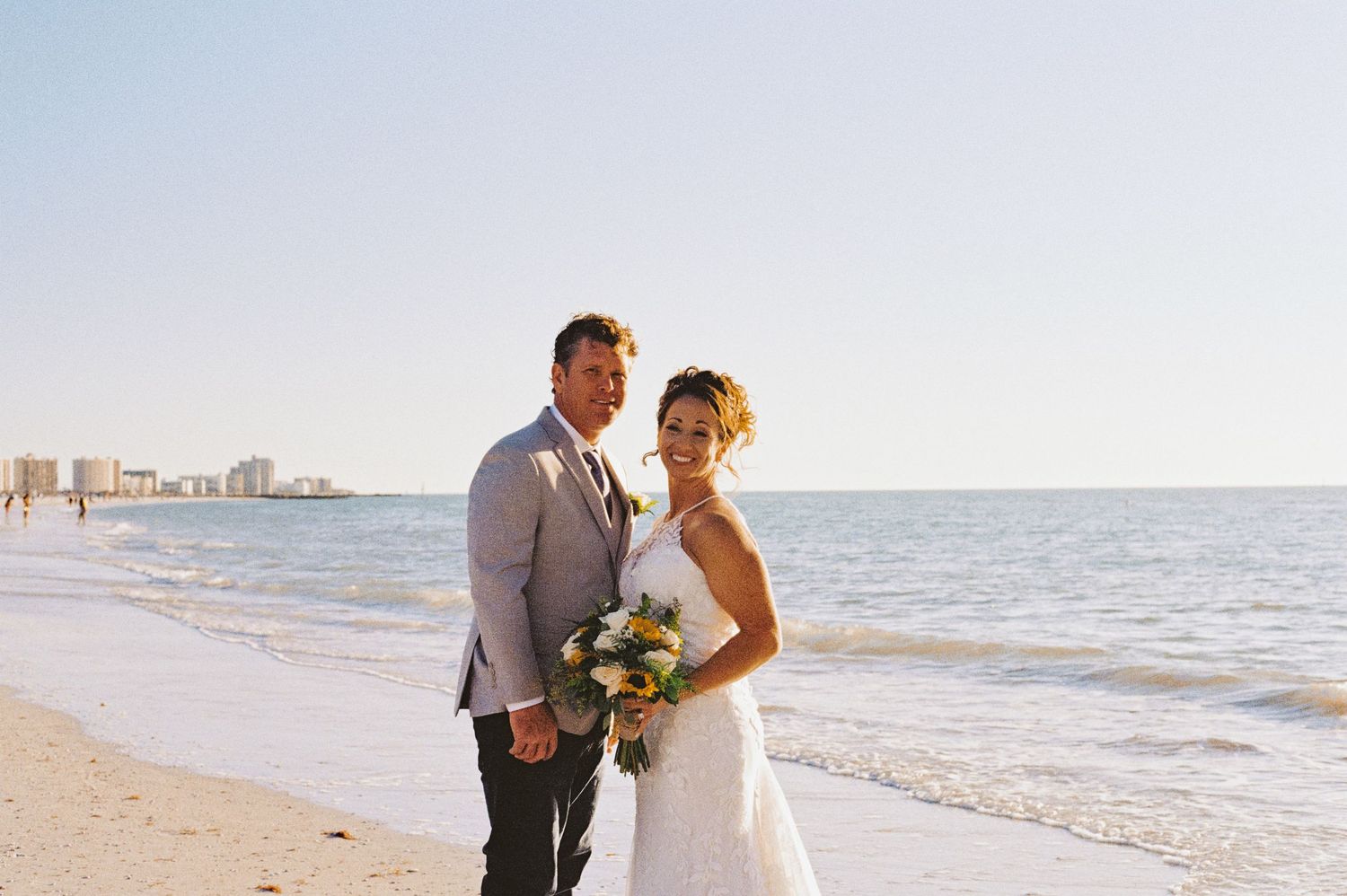 Bride and groom posing together on sandy beach with ocean and city skyline in background.