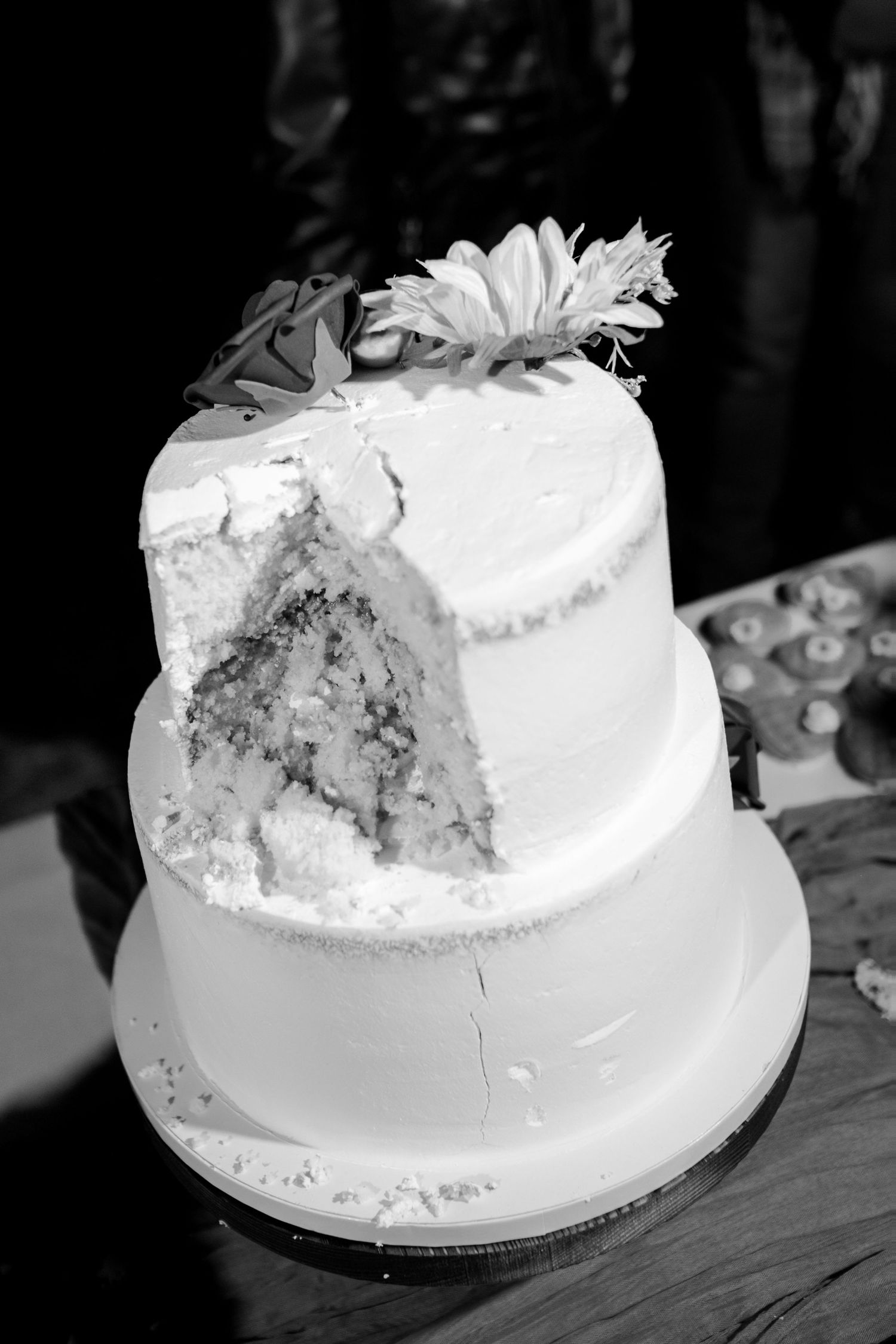 Black and white photo of a two-tier white wedding cake with floral topper, partially sliced.