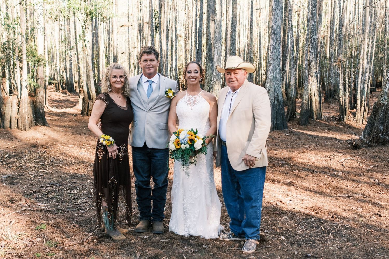 Bride and groom pose with two family members in a pine forest setting on wedding day.