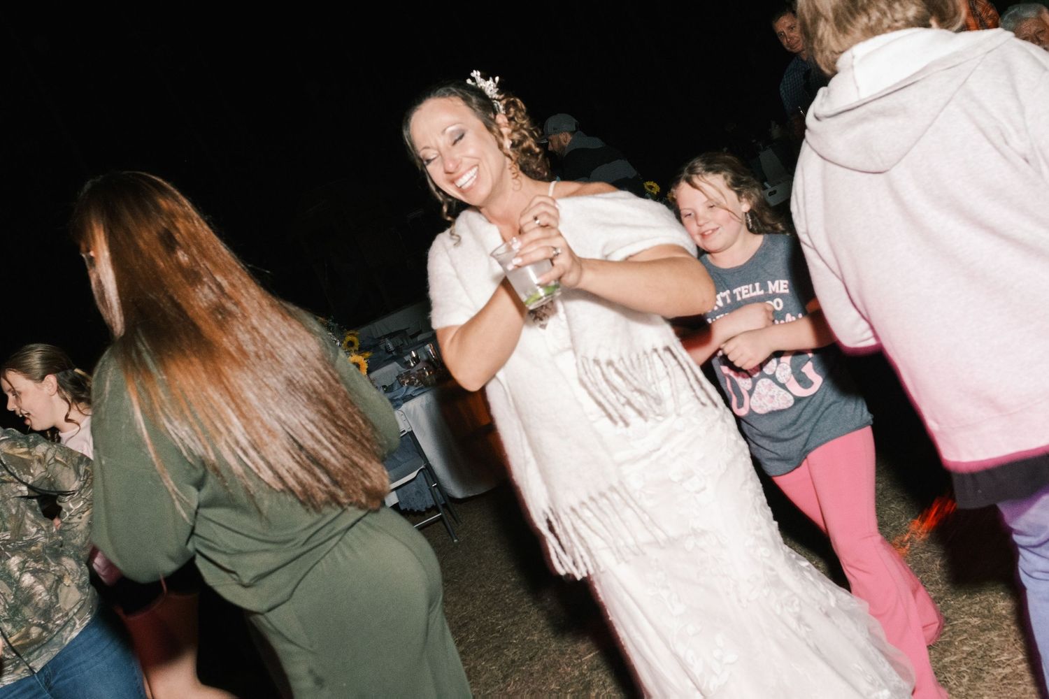 Bride in white lace dress dances joyfully with guests at outdoor evening wedding celebration.
