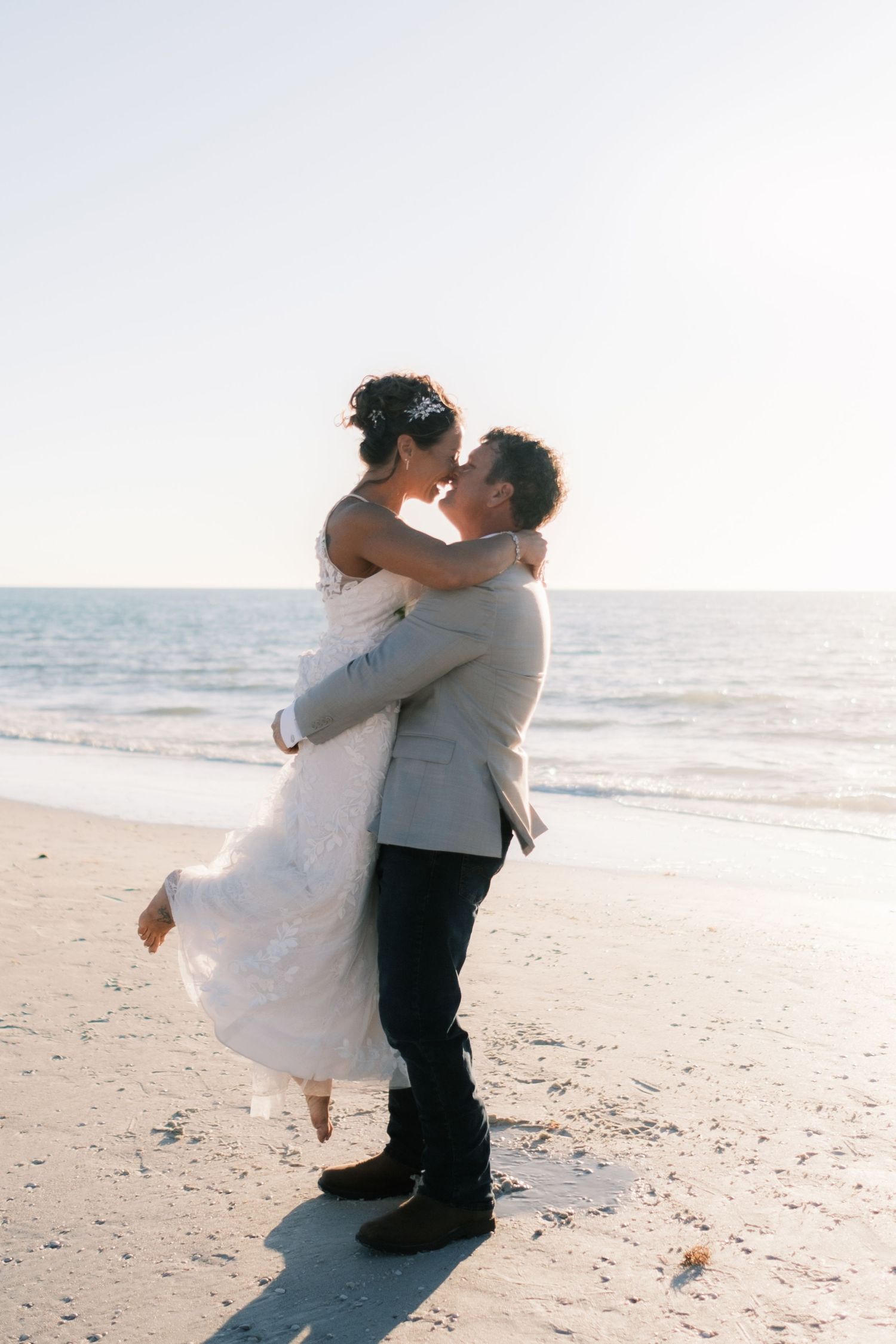 Groom lifts bride in lace wedding dress for a romantic kiss on a sunny beach shoreline.