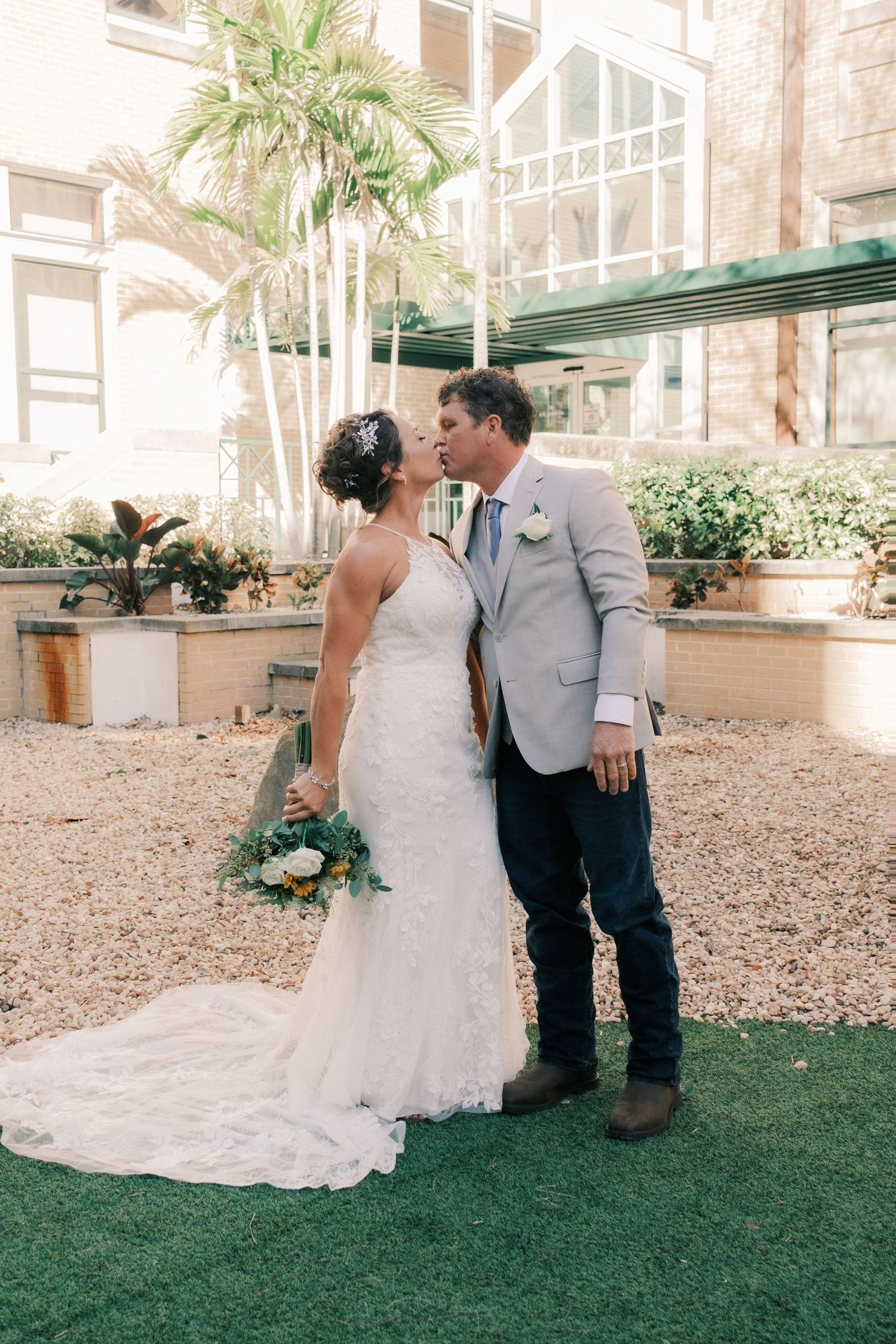Bride and groom sharing a kiss in courtyard with palm trees, bride holding bouquet.