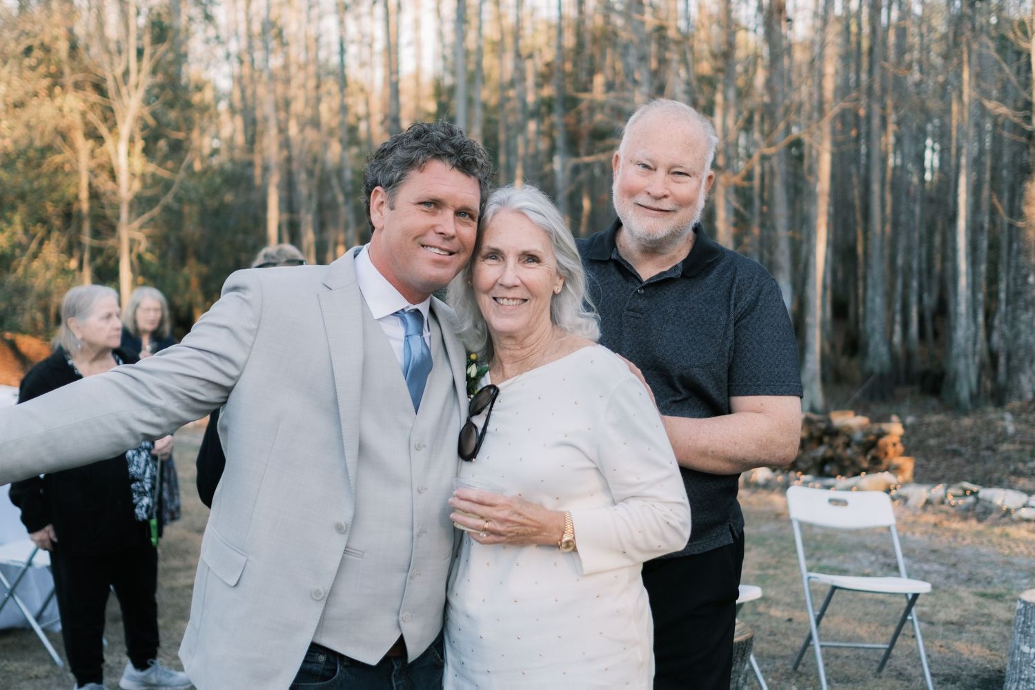 Three wedding guests smiling and posing together outdoors at a woodland wedding reception during golden hour.