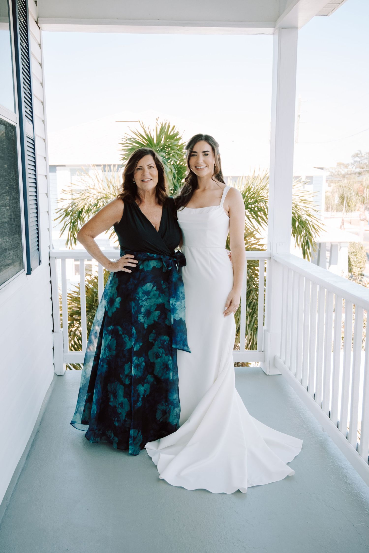Wedding portraits on a white porch with blue-green dress and white gown against coastal backdrop.