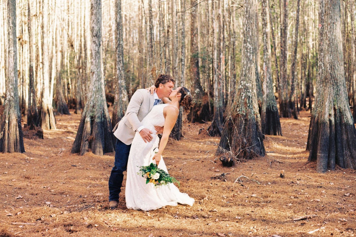 Groom dips bride for a romantic kiss among tall trees in a sunlit forest wedding portrait.