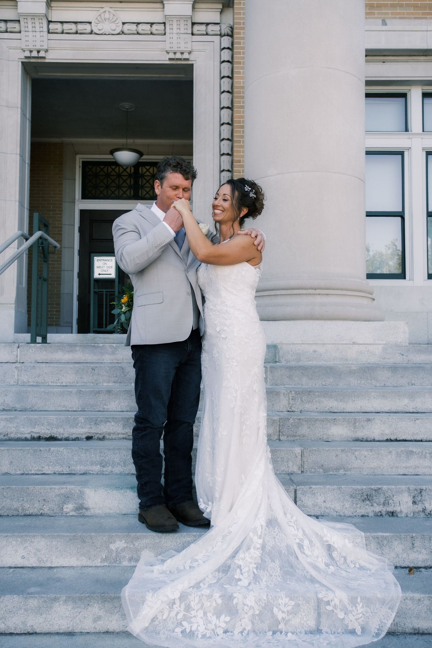 Groom kissing bride's hand as she smiles on steps of grand stone building in lace wedding gown.