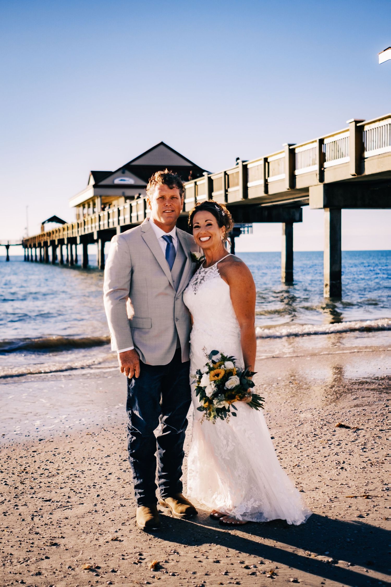 Smiling bride and groom posing on beach with pier in background.