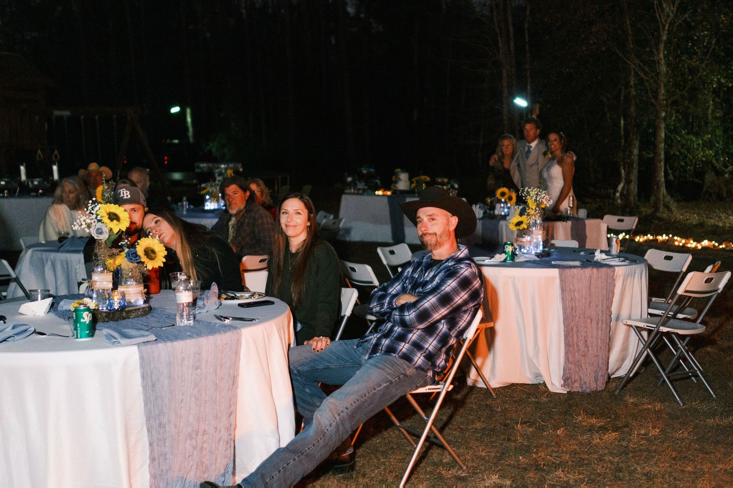 Guests seated at decorated round tables with sunflowers at an outdoor evening wedding reception.