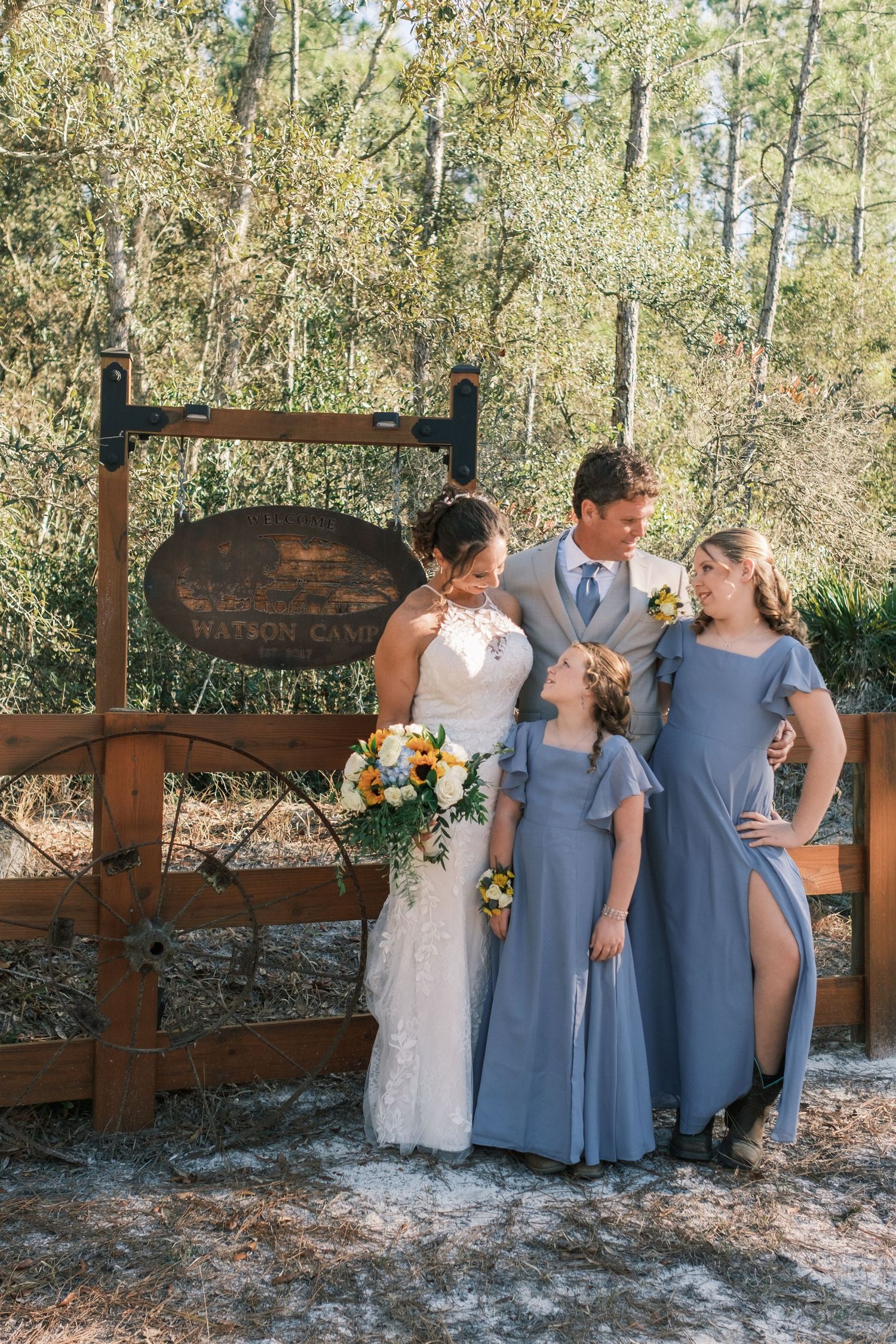 Wedding party with bride, groom, and two girls in blue dresses posing by a rustic wooden fence and sign outdoors.