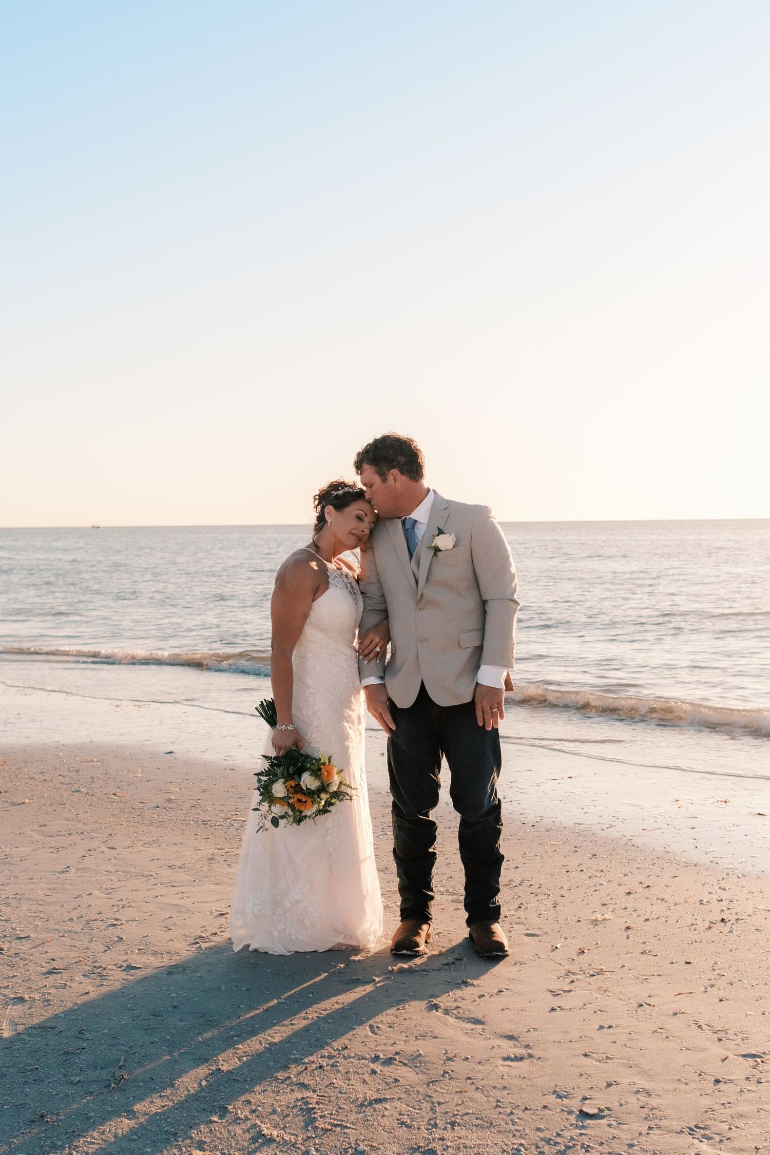 Bride and groom share a tender kiss on a sandy beach at sunset, bride holding colorful bouquet.