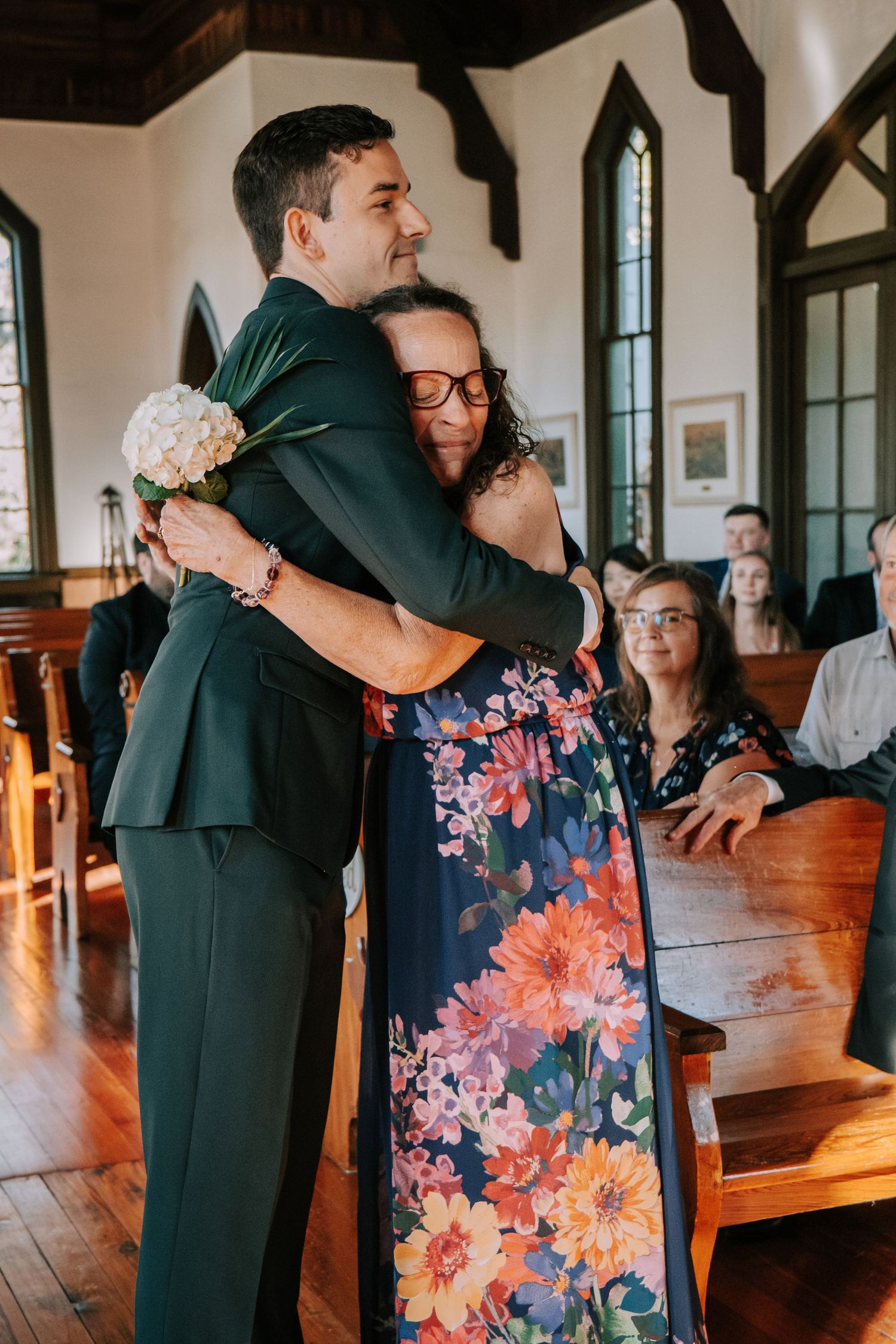 Emotional embraces between wedding attendees in a church with wooden pews and floral dress details.