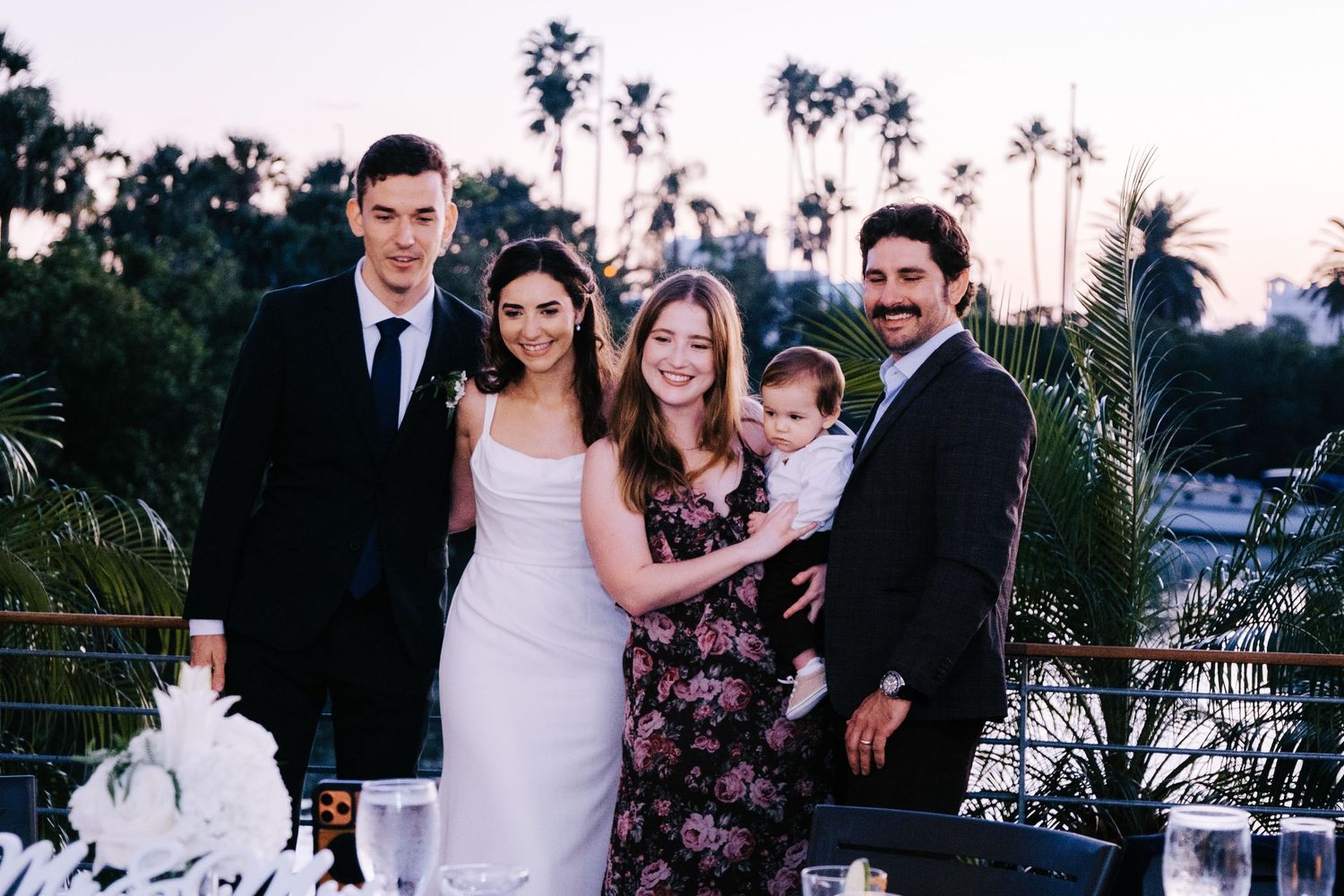 Wedding party gathering outdoors at dusk with palm trees silhouetted in the background.