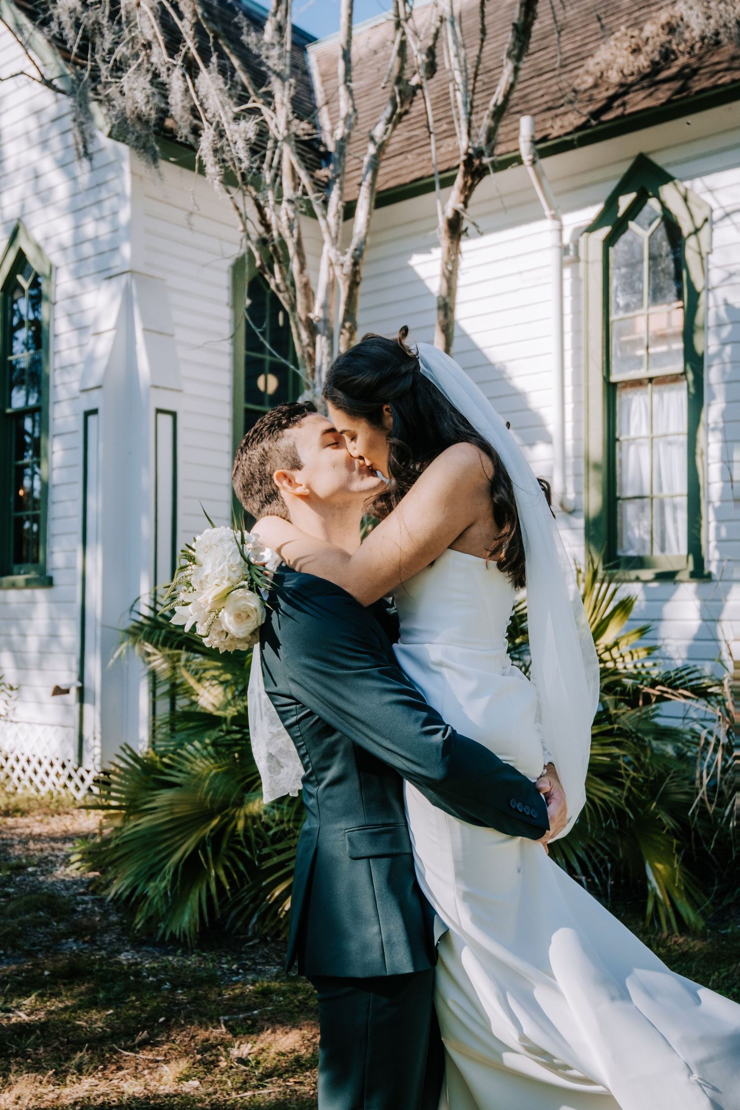 Romantic embrace between newlyweds outside white church with gothic windows.
