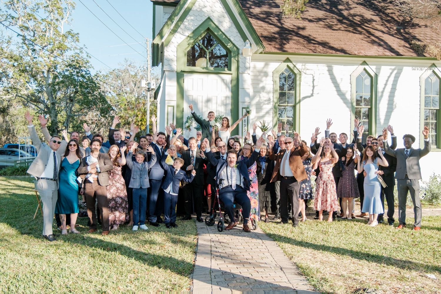 Wedding guests gather for a group photo outside a white chapel, raising their arms in celebration.