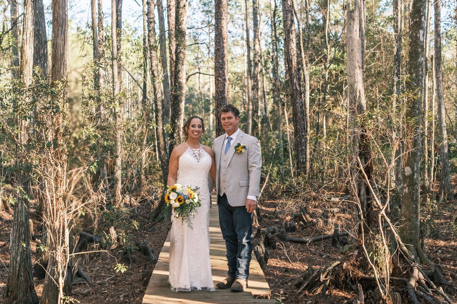 Bride in white lace gown and groom in tan suit stand together on wooden path in sunlit pine forest.