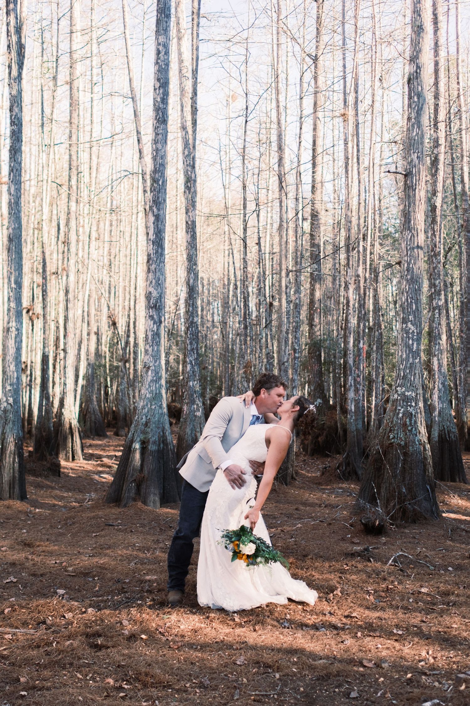 Groom dips bride for a kiss among tall bare trees in a sunlit forest during their wedding day.