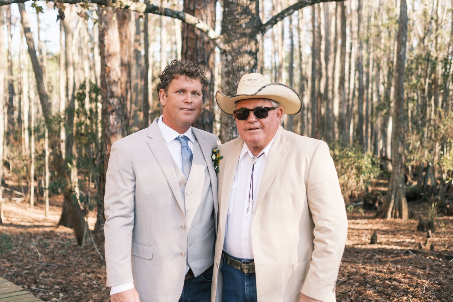 Groom in tan suit poses with an older man in a cowboy hat in a sunny woodland setting.