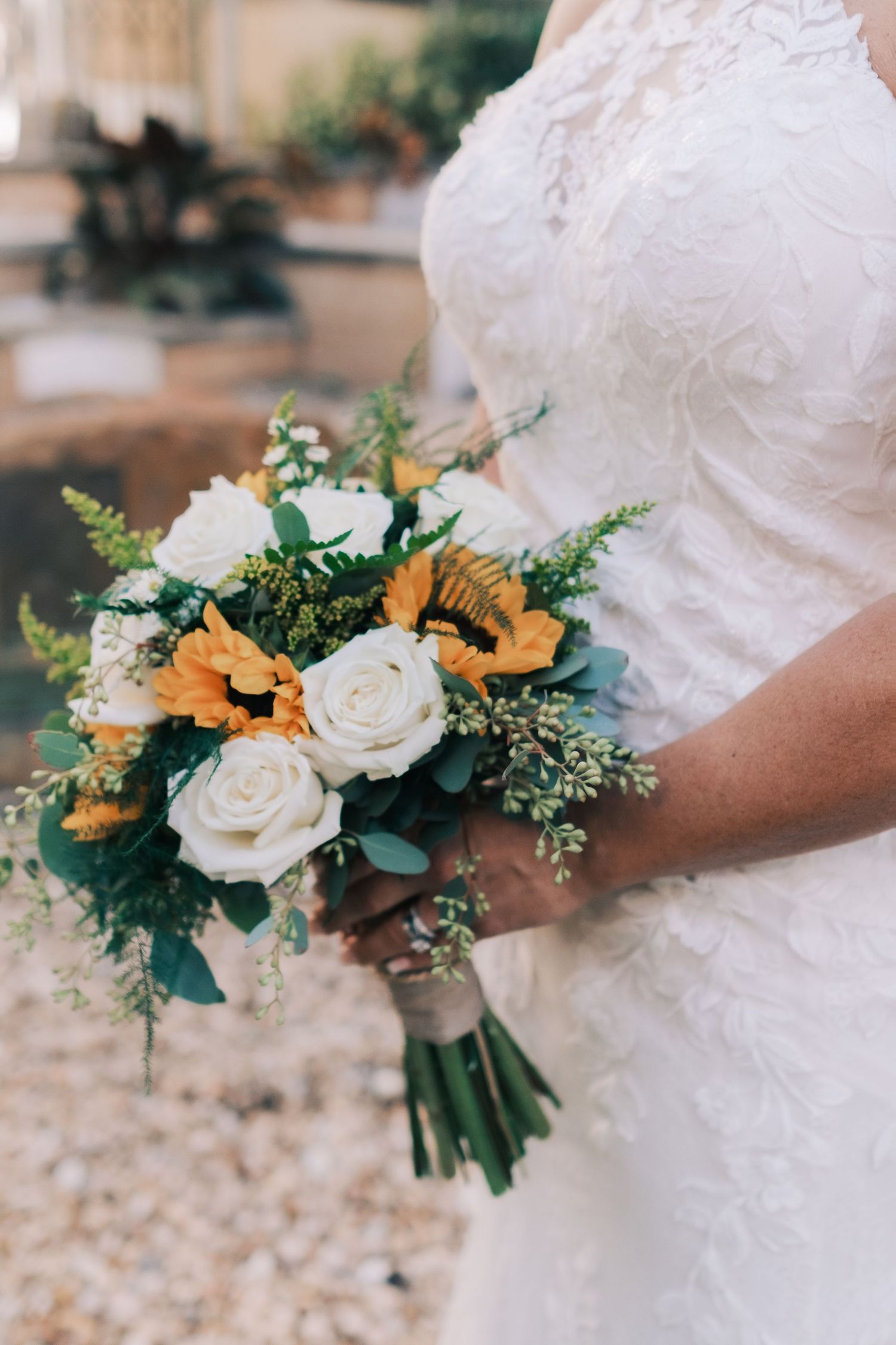 Bride holds a stunning bouquet of white roses and yellow sunflowers in a lace wedding dress.