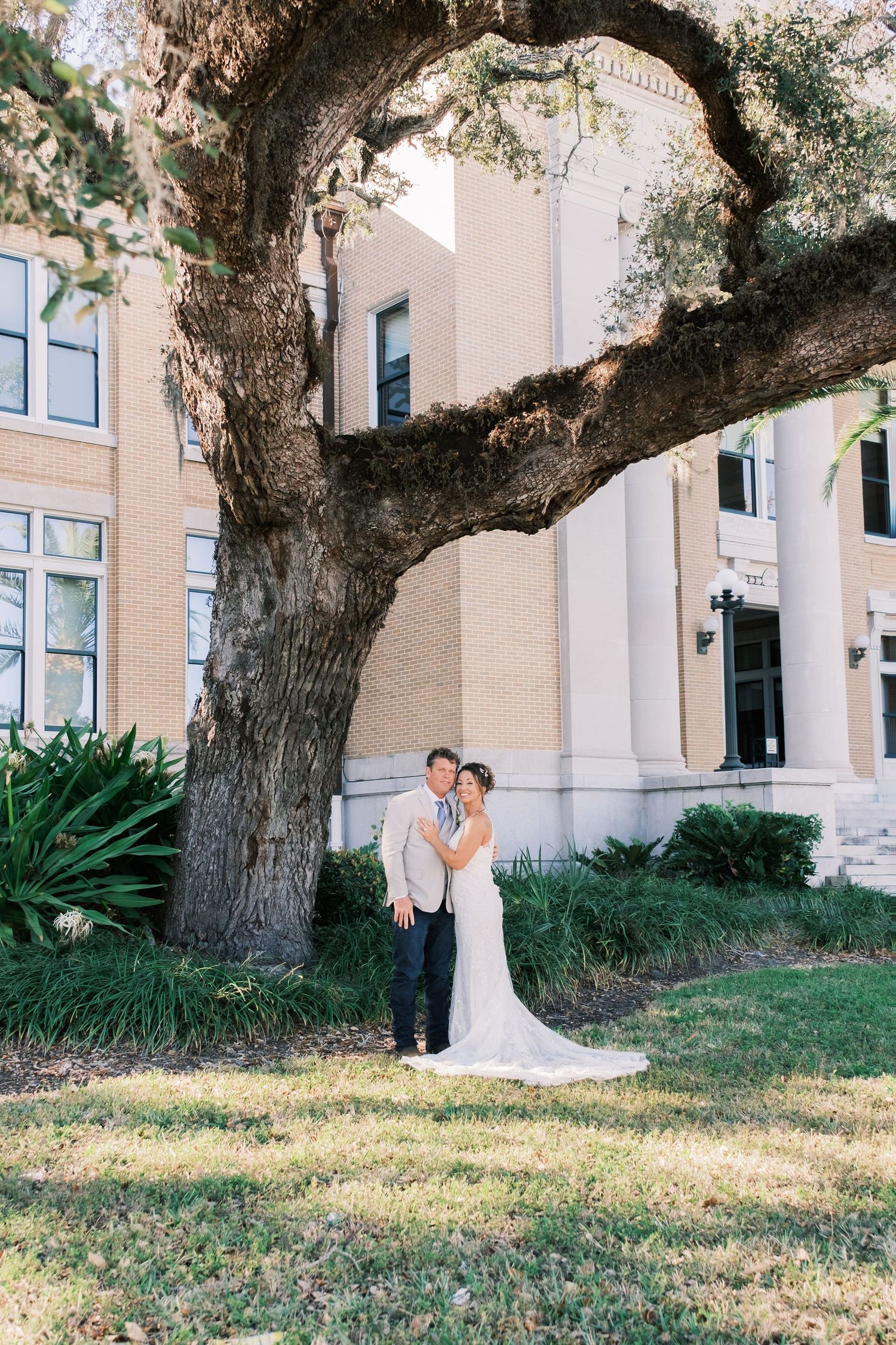 Bride and groom pose romantically under a sprawling oak tree beside an elegant brick building on their wedding day.