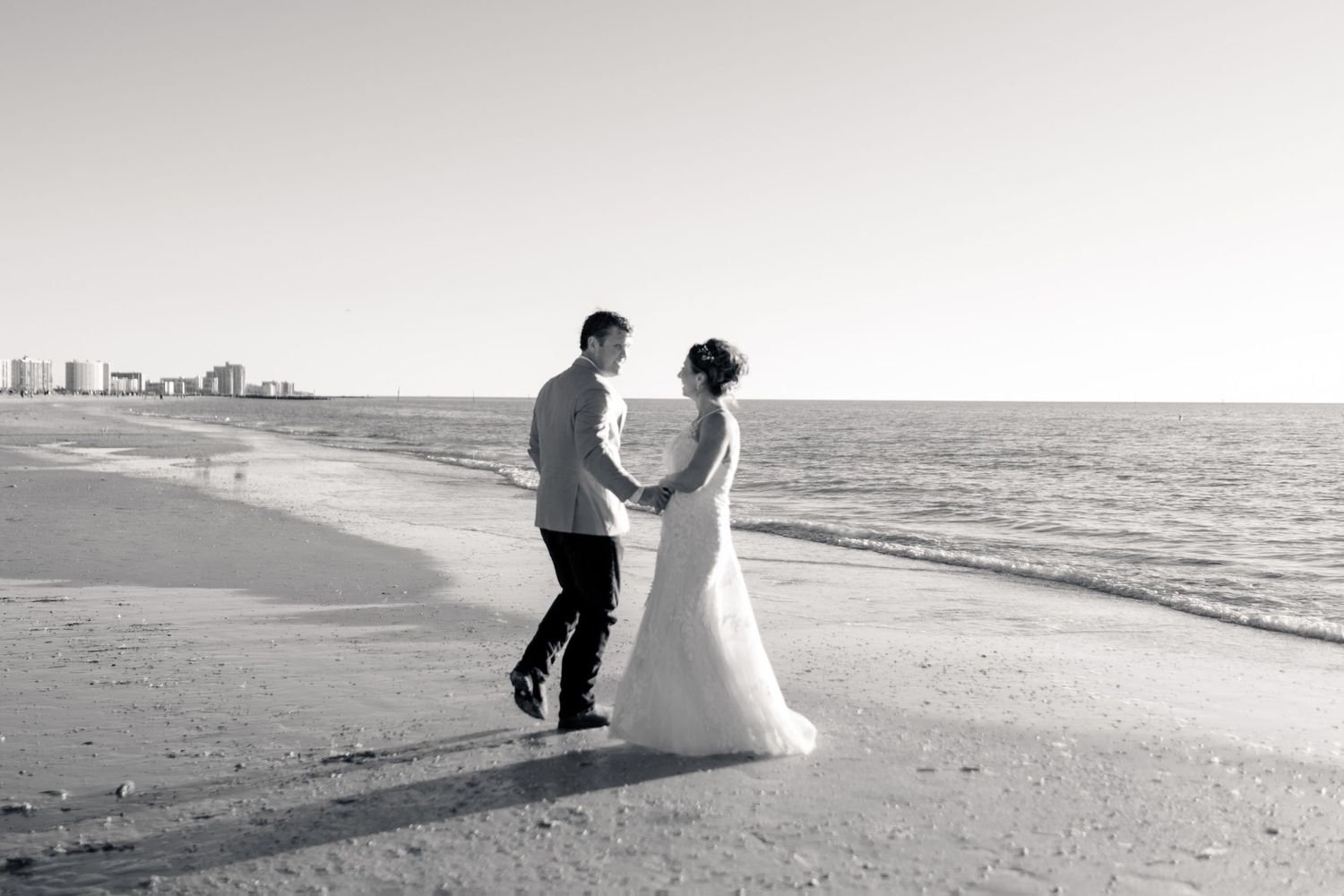 Black and white photo of bride and groom holding hands walking along ocean shoreline on wedding day.