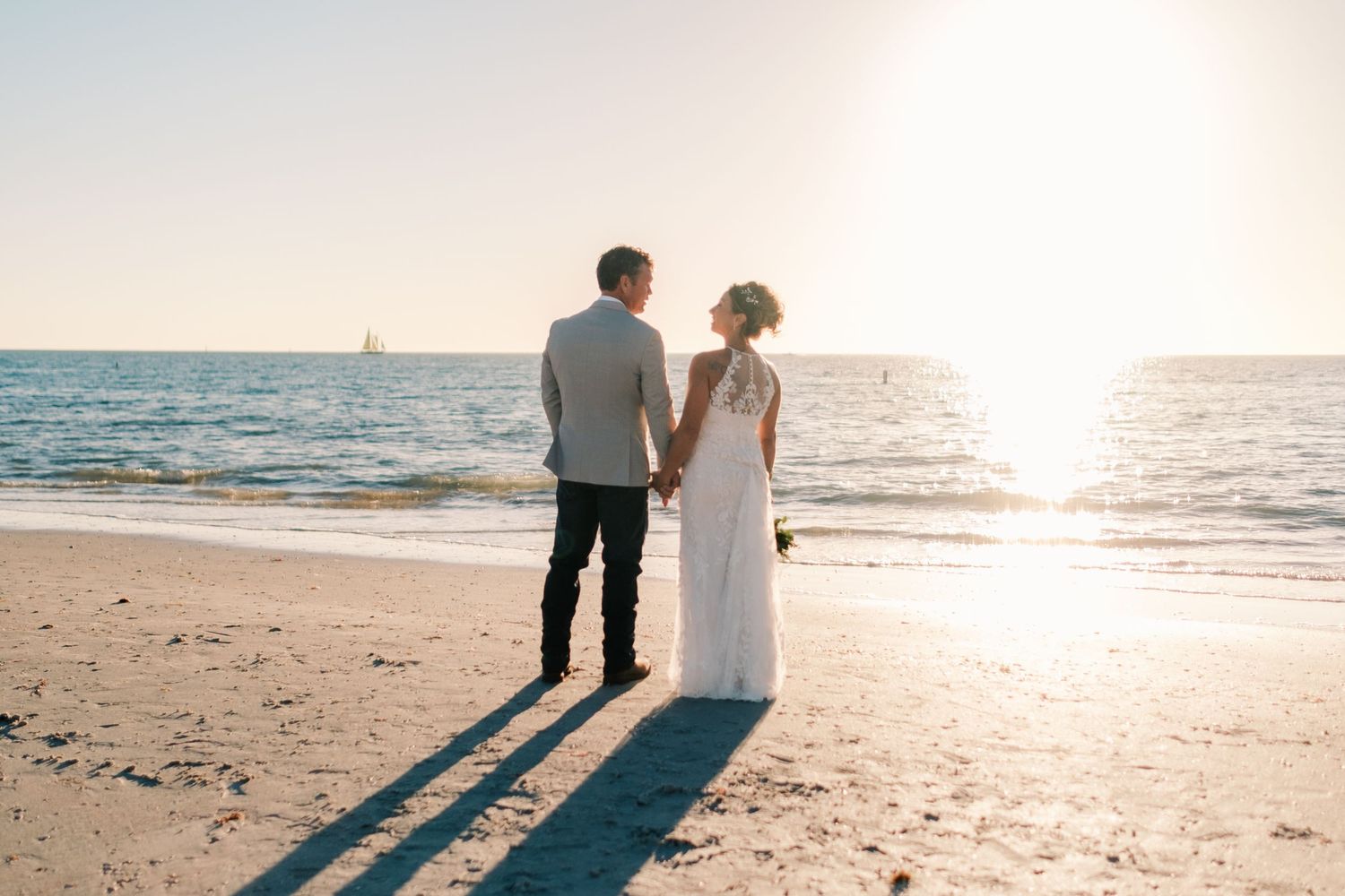 Bride and groom holding hands facing ocean at sunset on sandy beach, casting long shadows.