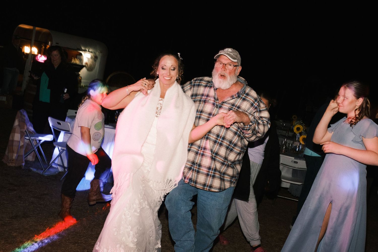 Bride in white gown dances with older man in plaid shirt at lively outdoor wedding reception.