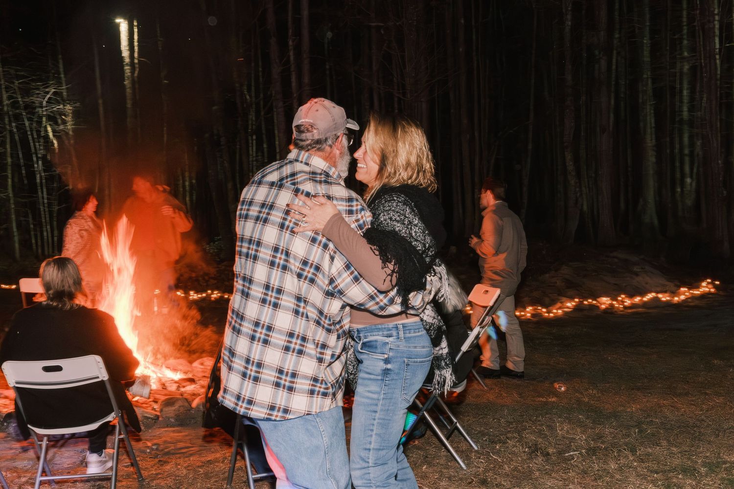 Couple embracing romantically in front of a glowing bonfire at an outdoor nighttime wedding reception.