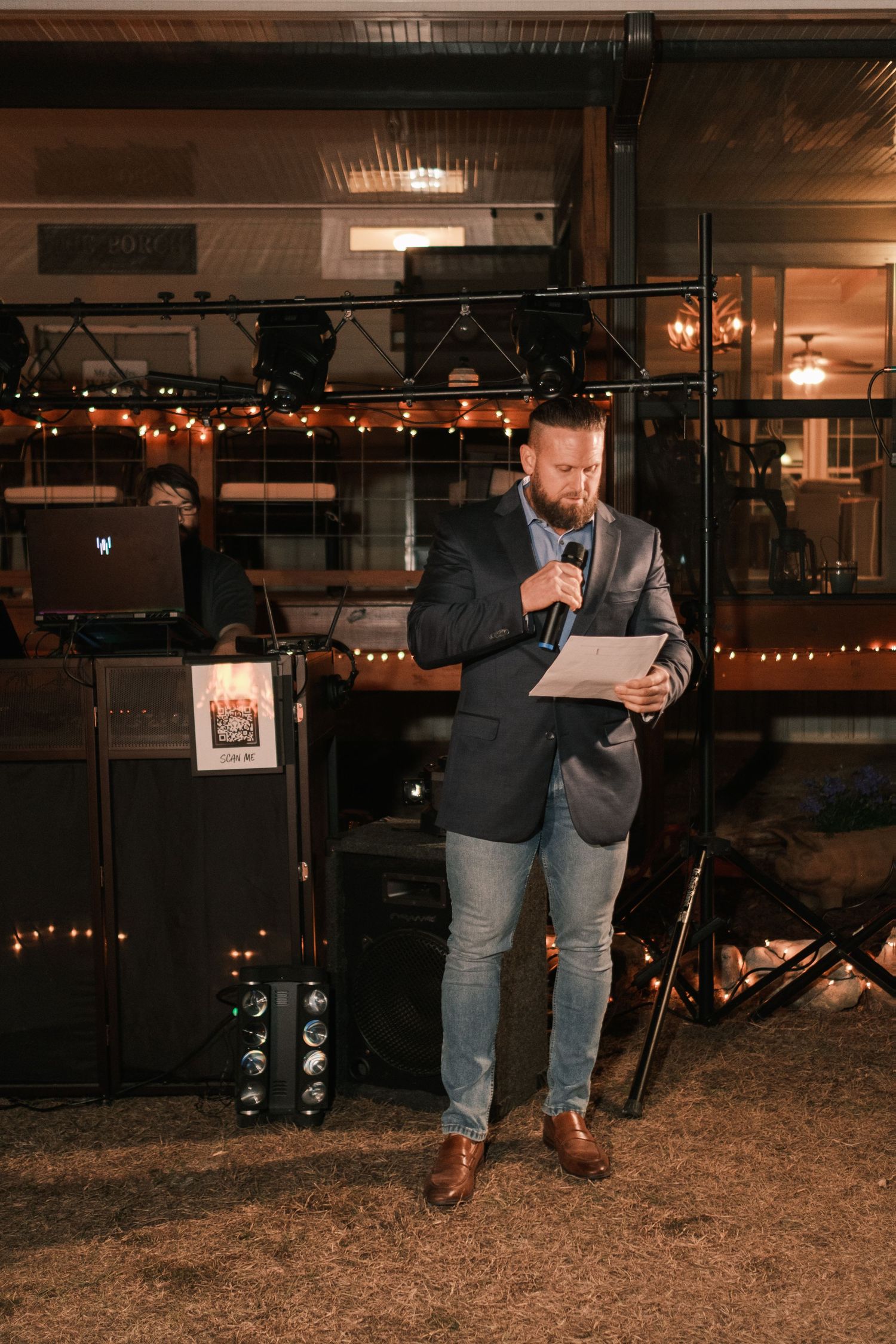Man in navy blazer delivers a speech holding a microphone and papers at a wedding reception with string lights.