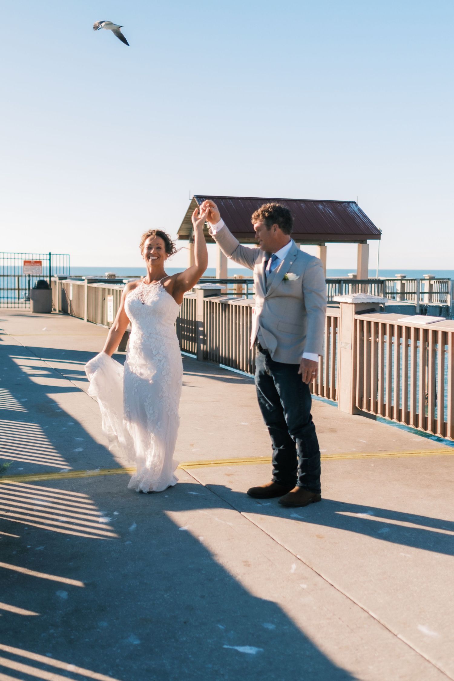 Groom twirls bride in white lace gown on sunny pier with seagull overhead.