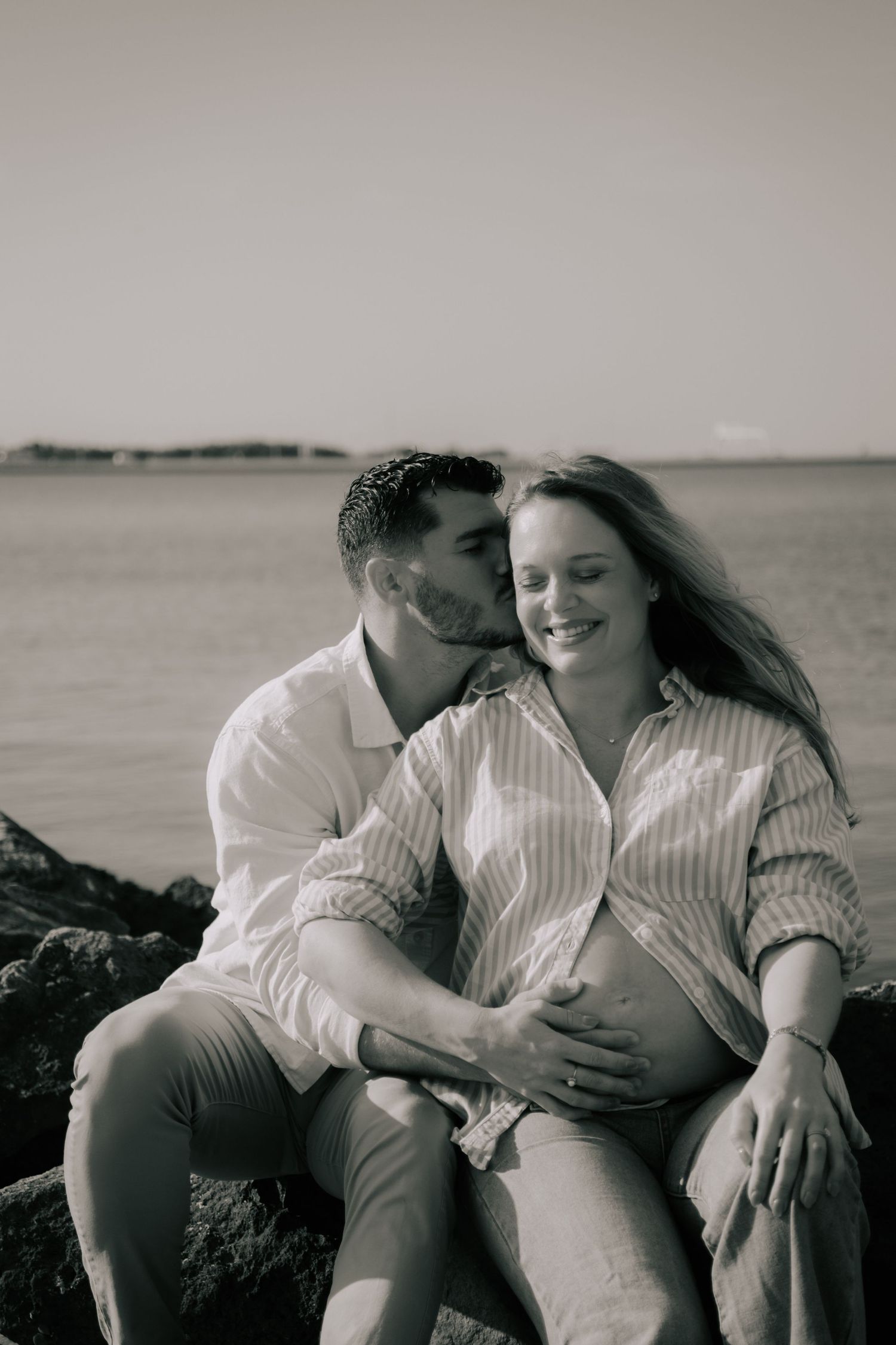 A romantic maternity photoshoot at the beach shows a couple sitting on rocks in black and white.