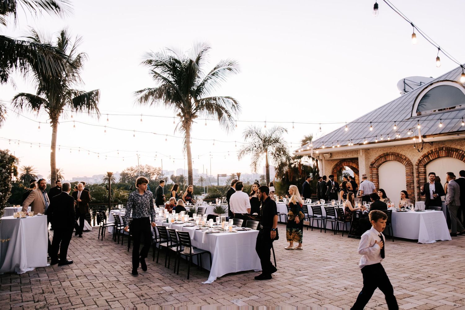 Evening wedding reception setup with string lights and palm trees on beachfront patio.