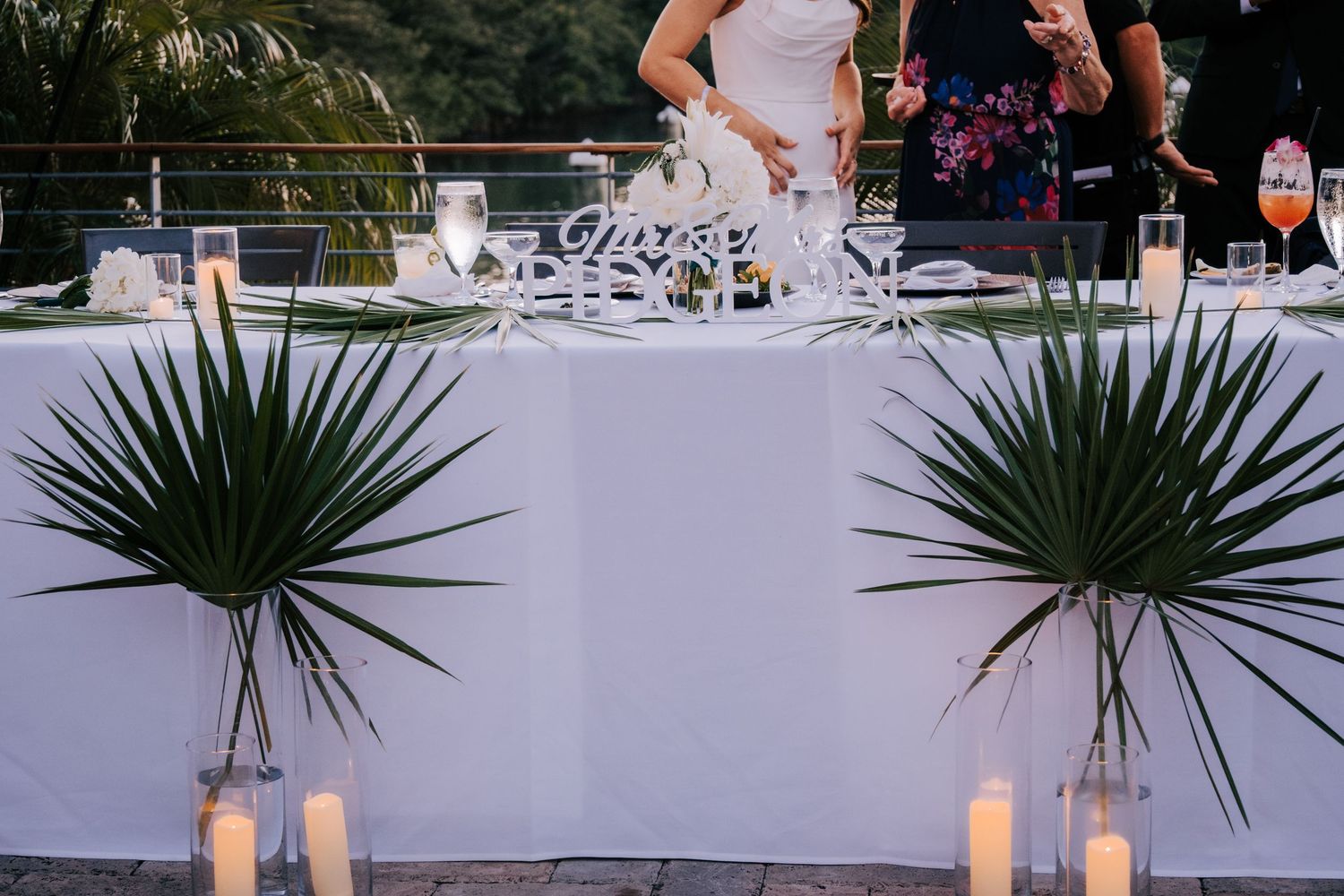 Elegant outdoor wedding reception setup with white tablecloths and tropical palm decorations at sunset.