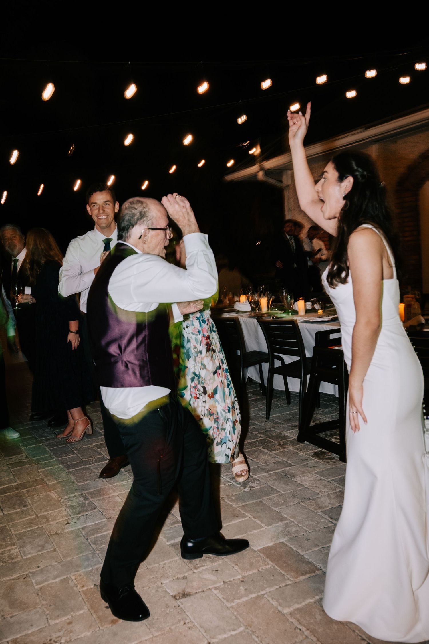 Wedding guests dancing and celebrating under string lights on a stone patio at night.