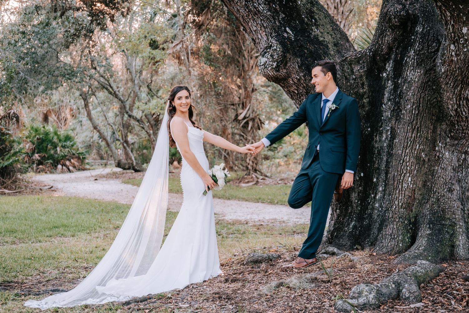 Bride in flowing veil holds groom's hand under massive oak tree.