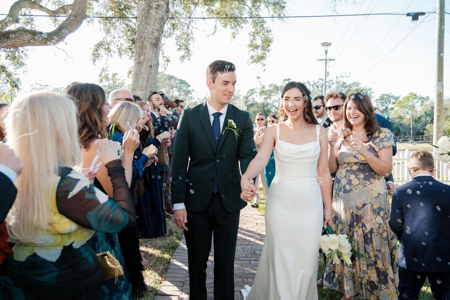 Outdoor wedding reception with guests mingling under trees and string lights.