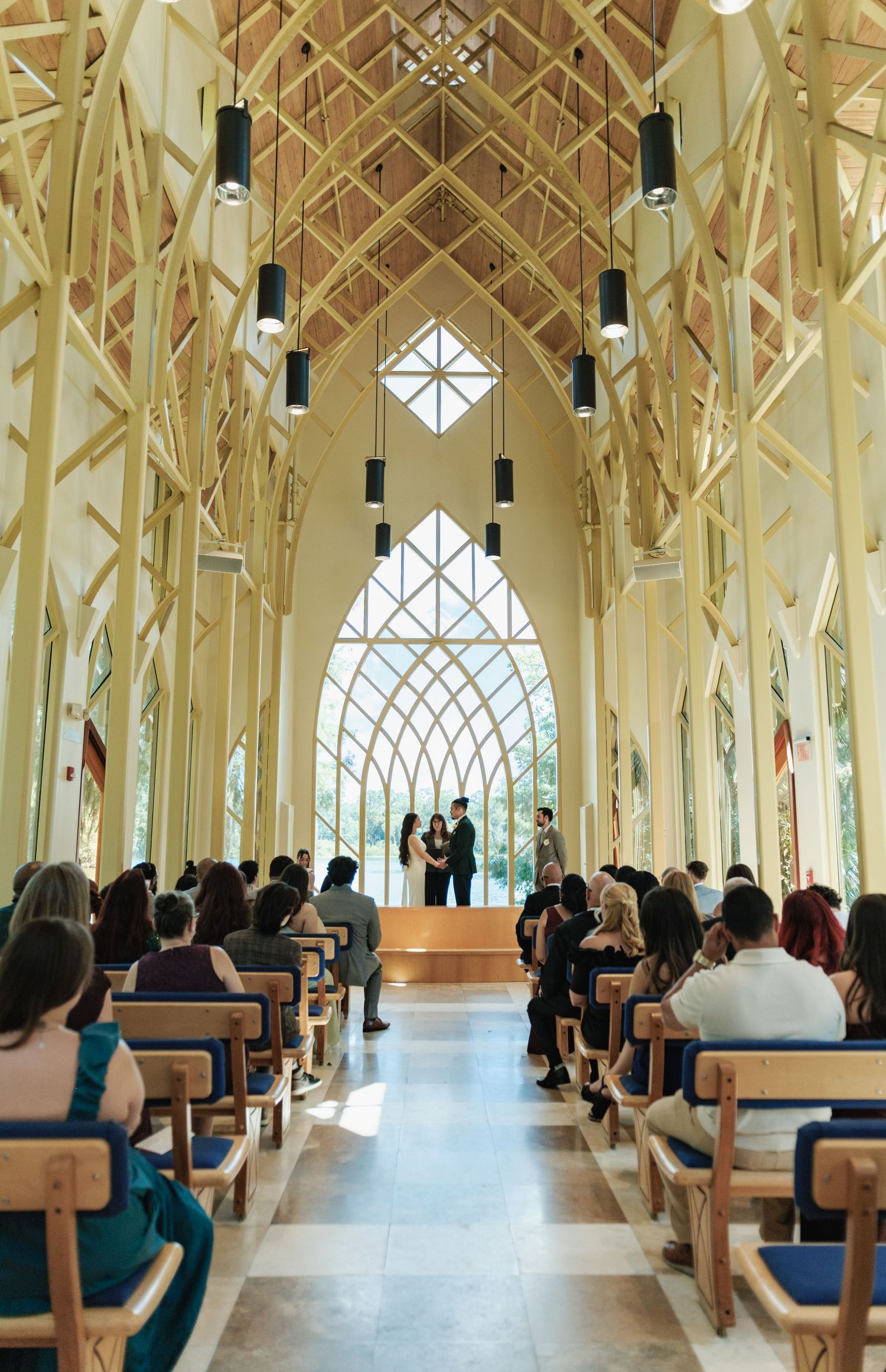 Modern chapel interior with wooden beams and geometric windows filled with wedding guests.