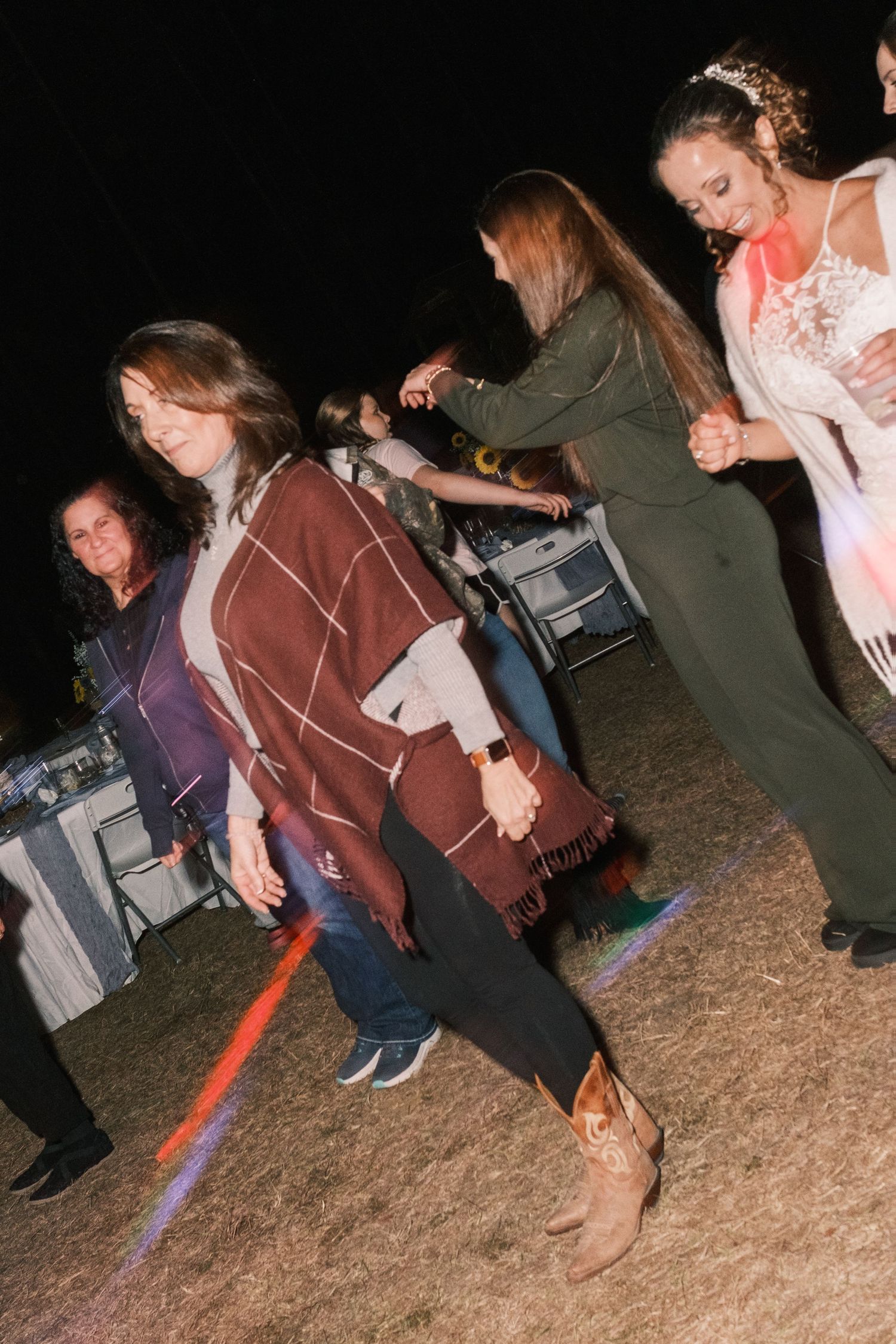 Women line dancing at an outdoor wedding reception at night with colorful stage lights.