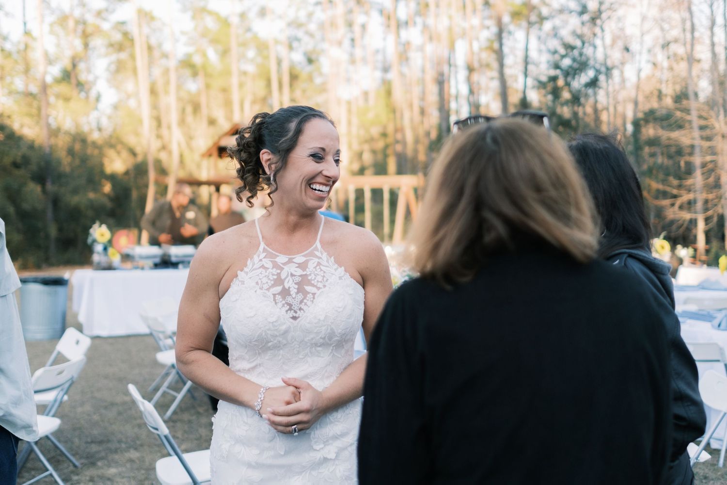 Smiling bride in a lace halter wedding dress chatting with a guest at an outdoor wedding reception.