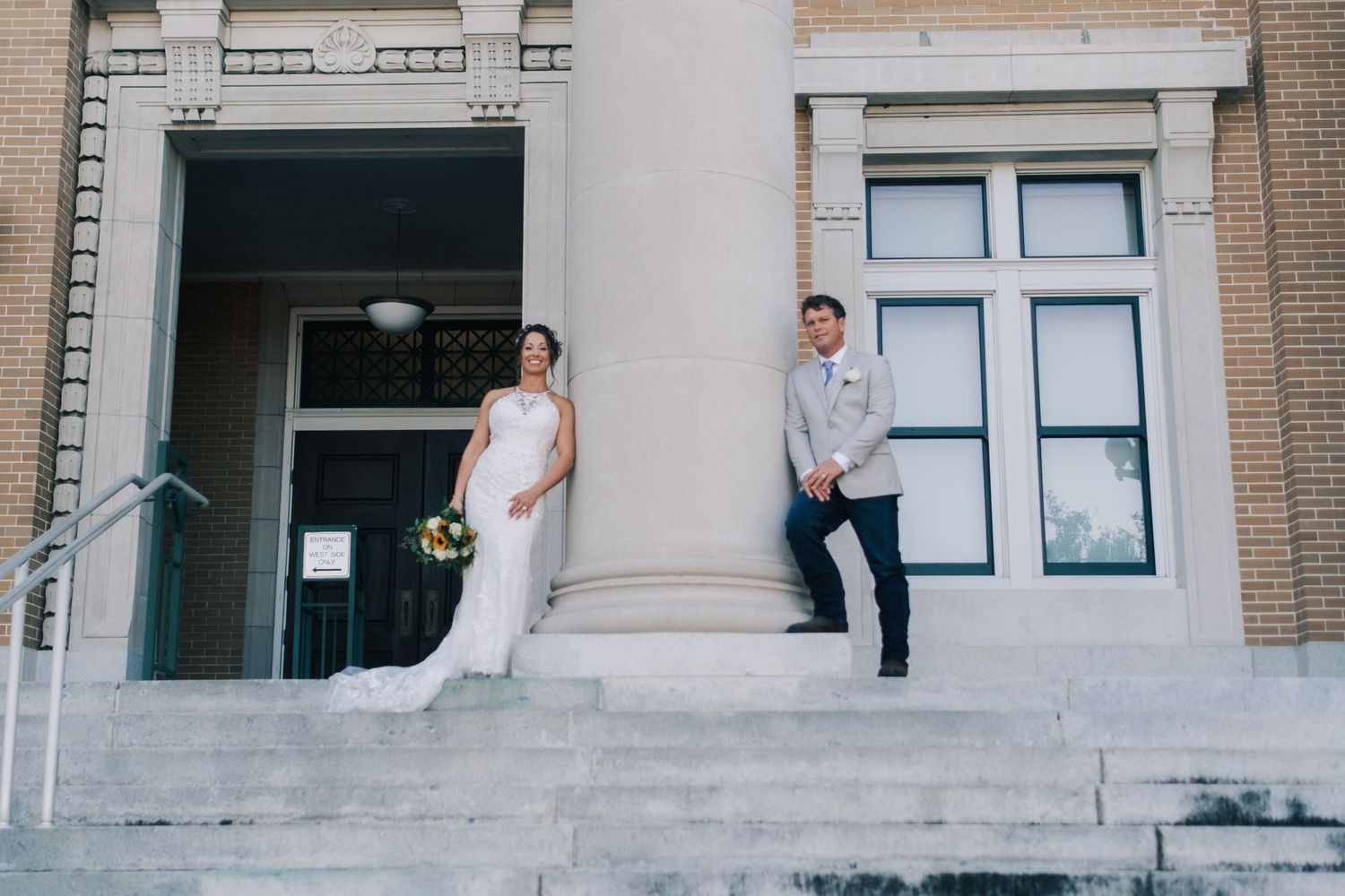 Wedding couple stands apart on grand stone steps of classic brick building, bride holding bouquet.