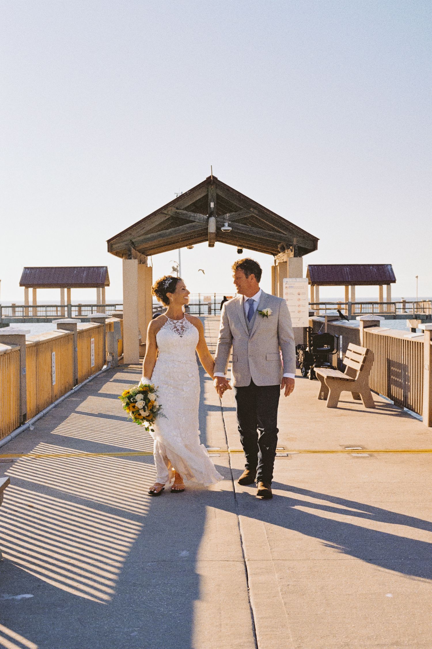 Bride and groom walk hand-in-hand along a sunny waterfront pier, bride holding a vibrant yellow bouquet.
