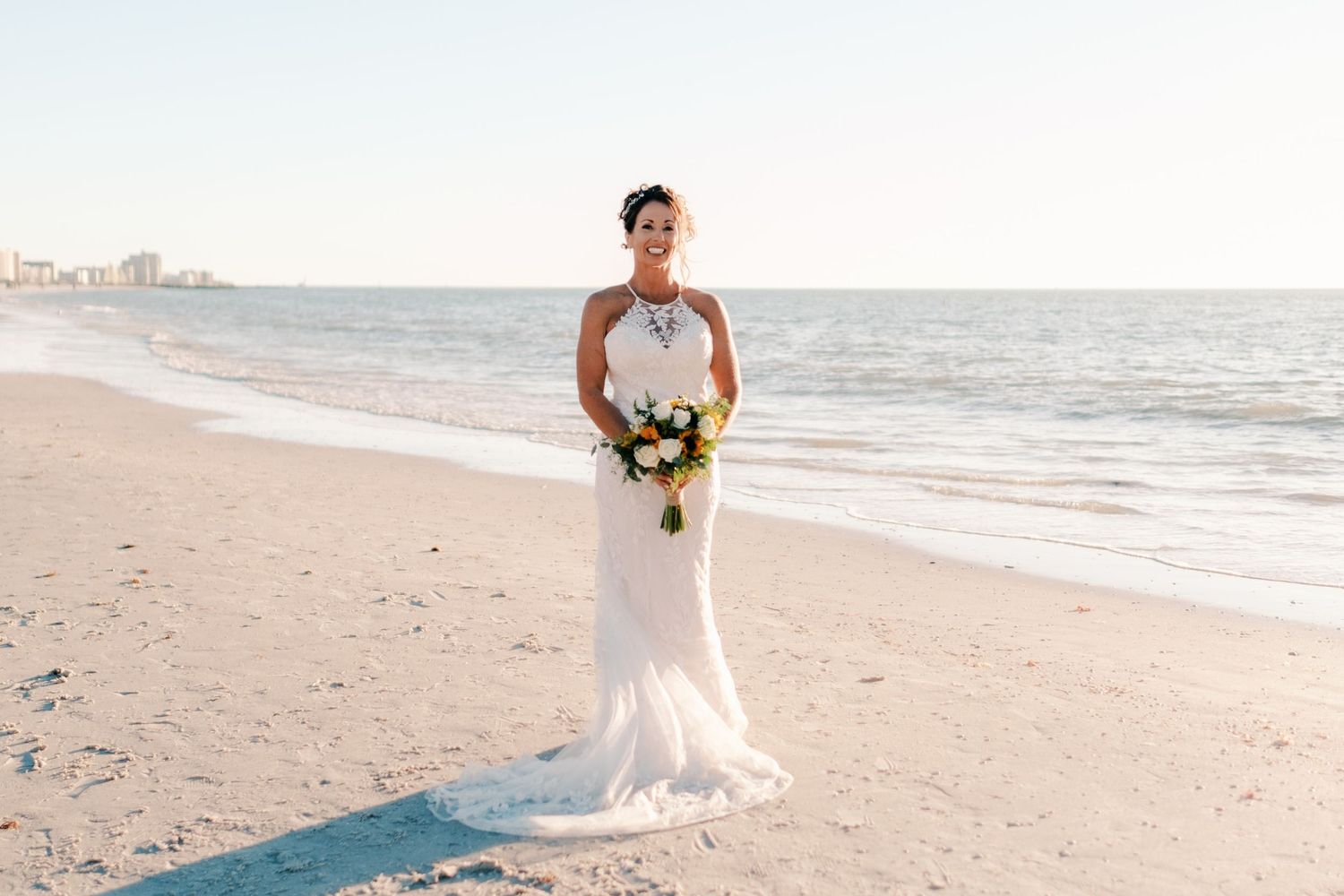 Bride in elegant halter lace gown holds colorful bouquet while posing on a sunny sandy beach.