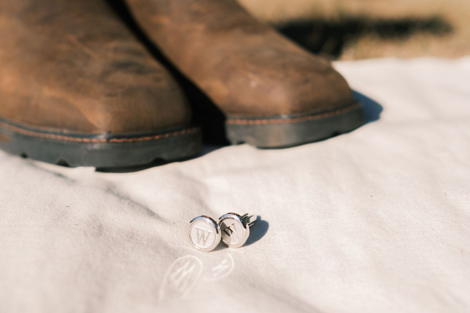 Brown leather cowboy boots with silver monogram cufflinks on white fabric for wedding day.